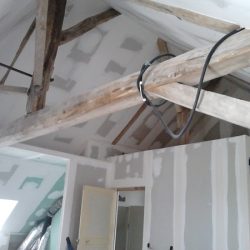 Room under construction with exposed wooden rafters, loose wires, and partially finished drywall on walls and ceiling on a worksite.