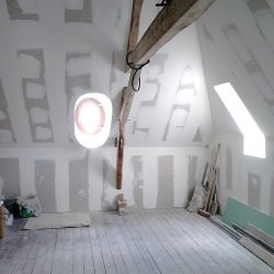 Attic under renovation with unfinished drywall, a wooden beam across the ceiling, and a round wall/window opening surrounded by light. Construction materials and tools are scattered on the floor and walls are paneled with drywall seams visible.