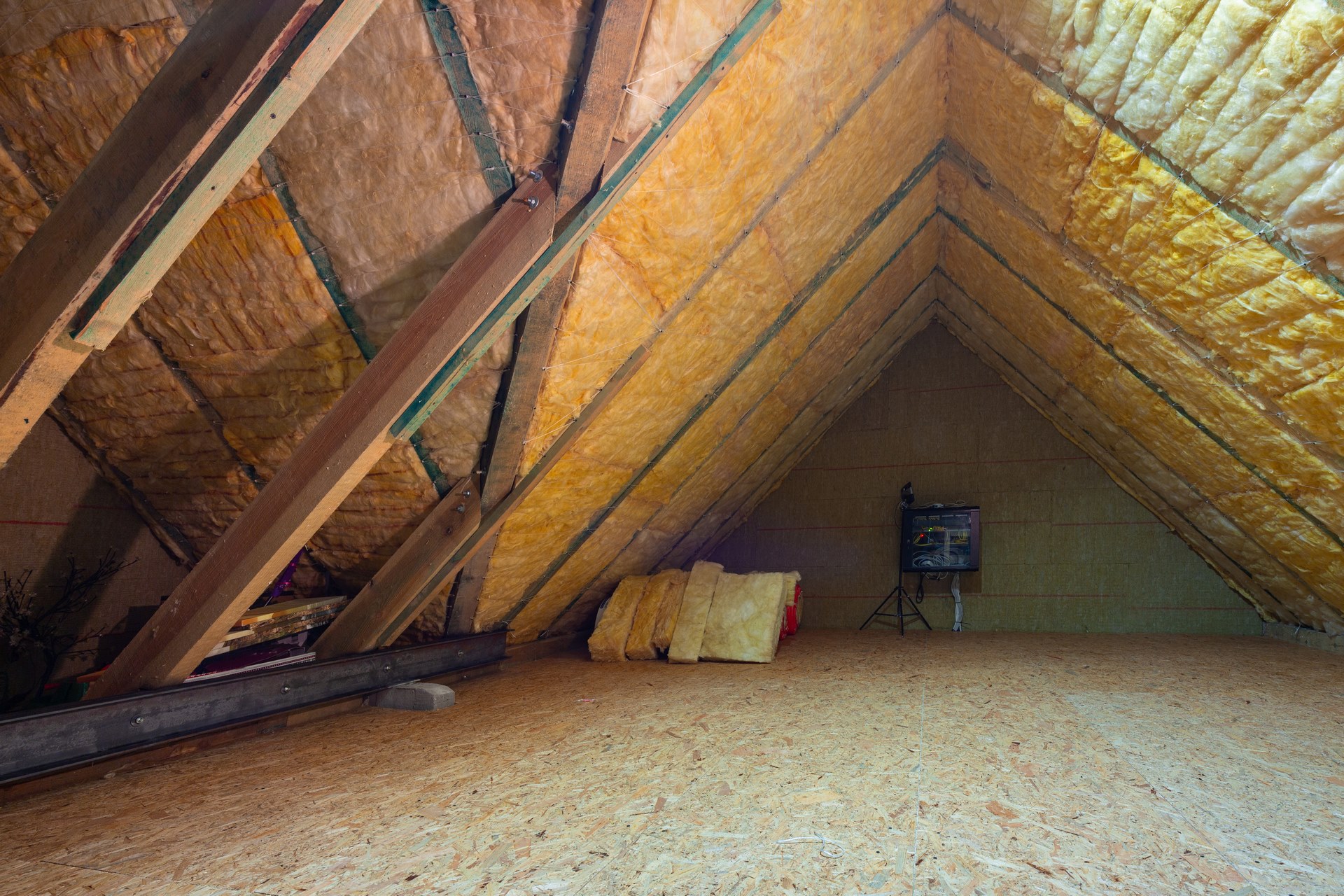 Attic space with exposed wooden rafters, yellow insulation, and unfinished plywood floor; construction materials and a small electrical panel in the back.