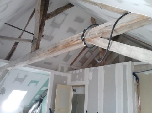 Room under construction with exposed wooden rafters, loose wires, and partially finished drywall on walls and ceiling on a worksite.