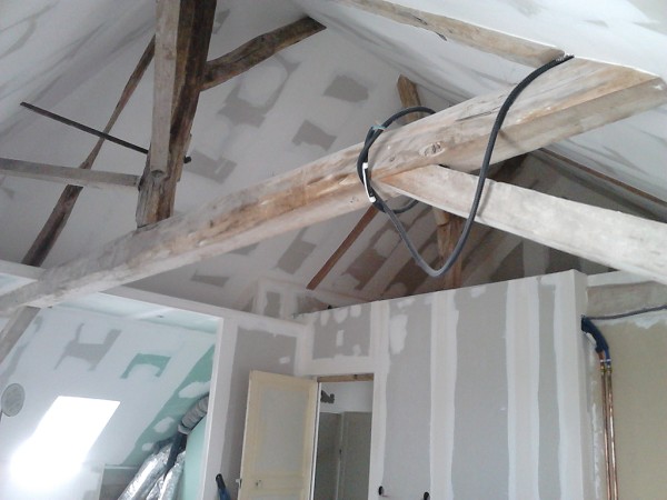 Room under construction with exposed wooden rafters, loose wires, and partially finished drywall on walls and ceiling on a worksite.