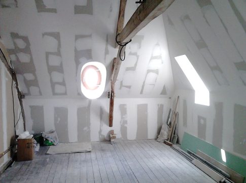 Attic under renovation with unfinished drywall, a wooden beam across the ceiling, and a round wall/window opening surrounded by light. Construction materials and tools are scattered on the floor and walls are paneled with drywall seams visible.