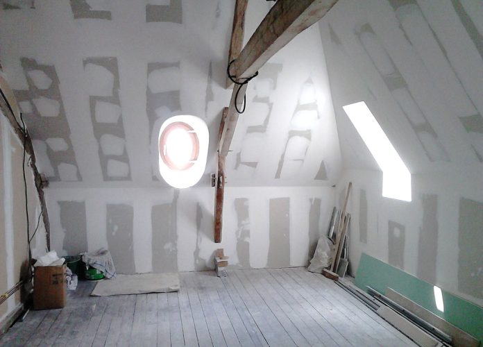 Attic under renovation with unfinished drywall, a wooden beam across the ceiling, and a round wall/window opening surrounded by light. Construction materials and tools are scattered on the floor and walls are paneled with drywall seams visible.