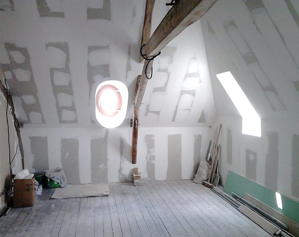 Attic under renovation with unfinished drywall, a wooden beam across the ceiling, and a round wall/window opening surrounded by light. Construction materials and tools are scattered on the floor and walls are paneled with drywall seams visible.