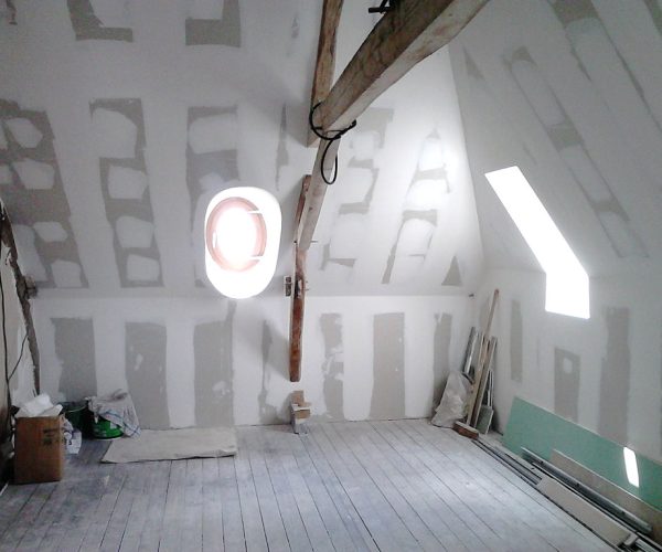 Attic under renovation with unfinished drywall, a wooden beam across the ceiling, and a round wall/window opening surrounded by light. Construction materials and tools are scattered on the floor and walls are paneled with drywall seams visible.